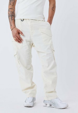 Pantaloni cargo crema con multiple tasche, fascia elastica in vita e vestibilità dritta, abbinati a sneakers grigio chiaro. Tessuto con una superficie liscia.