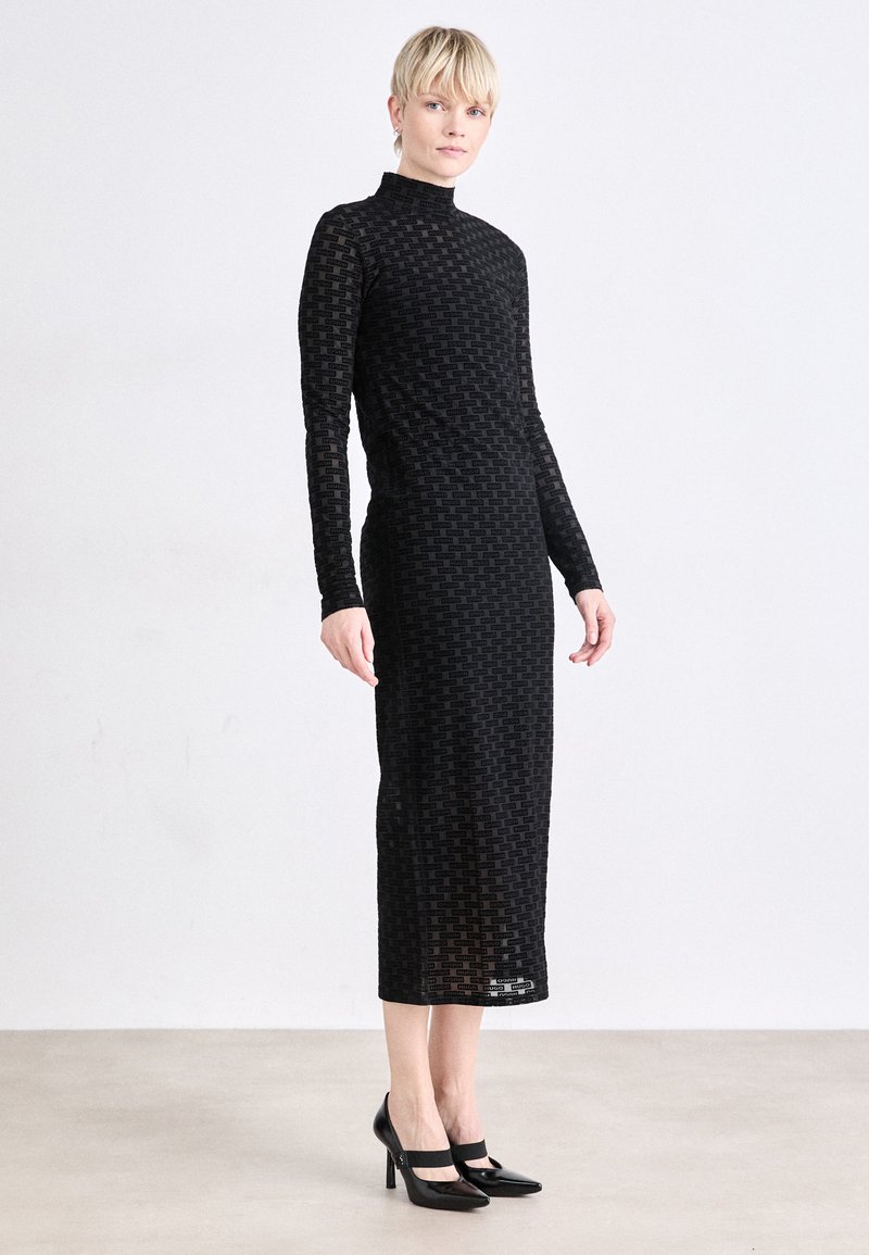 Black long-sleeve midi dress with a high neckline, featuring a patterned, sheer fabric. Paired with black high-heeled shoes.