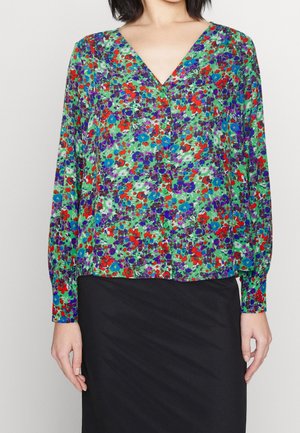 Vrouw die een groene V-hals blouse draagt met rood, paars en blauw bloemenprint en een zwarte rok, staand tegen een effen achtergrond.