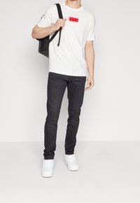 Valkoinen puuvillapaita, jossa on punainen graafinen kuva, yhdistettynä tummiin farkkuihin ja valkoisiin lenkkitossuihin. Malli pitää mustaa reppua toisen olkapään yli.