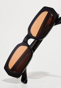Gafas de sol rectangulares negras con montura gruesa y brillante y lentes de color marrón. Presentan un sutil logotipo en el interior de la patilla.