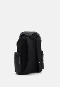 Svart ryggsäck med vadderade remmar, rutiga sidofickor med dragkedjor och en överdel med lock på vit bakgrund.