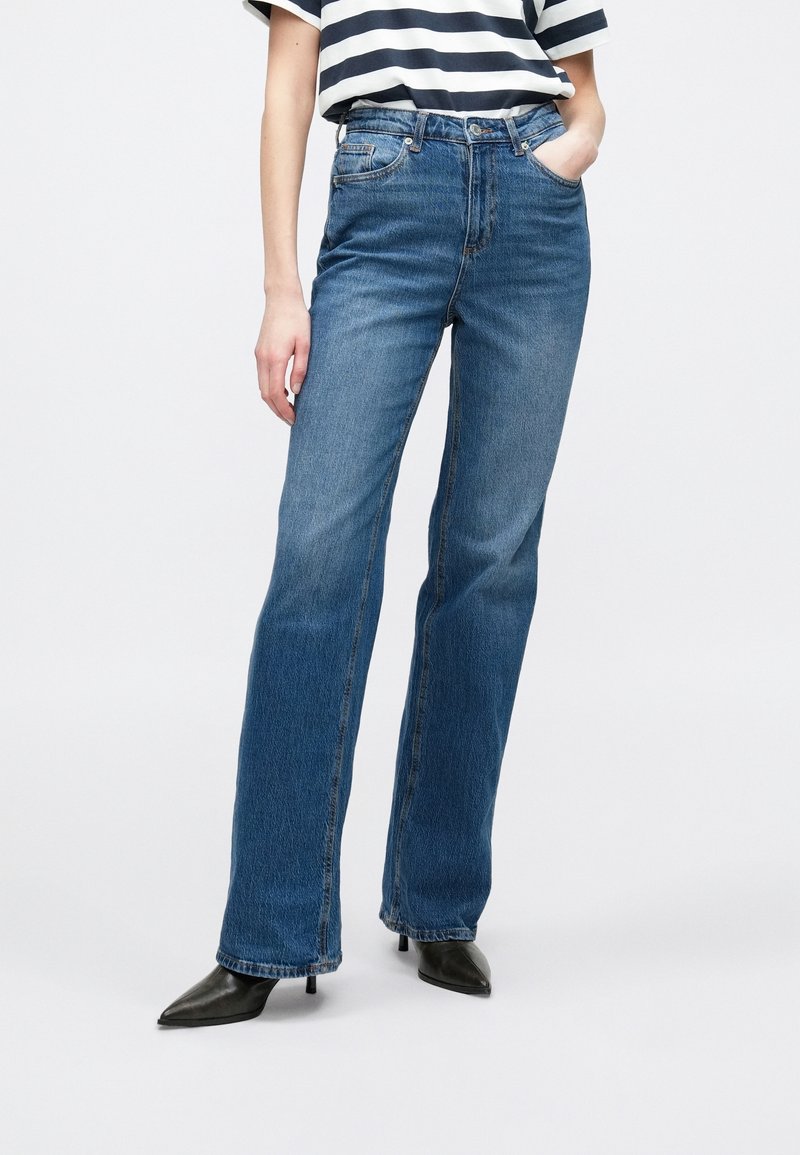 Personne portant un jean bleu taille haute à jambes droites, des talons noirs à bouts pointus et un chemisier à manches courtes rayé bleu marine et blanc.