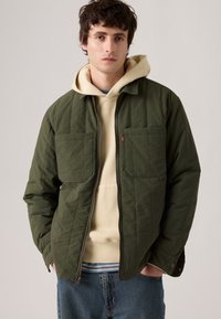 Veste matelassée olive avec deux poches poitrine, une fermeture éclair à l'avant, et un sweat à capuche beige en dessous, portée avec un jean bleu.