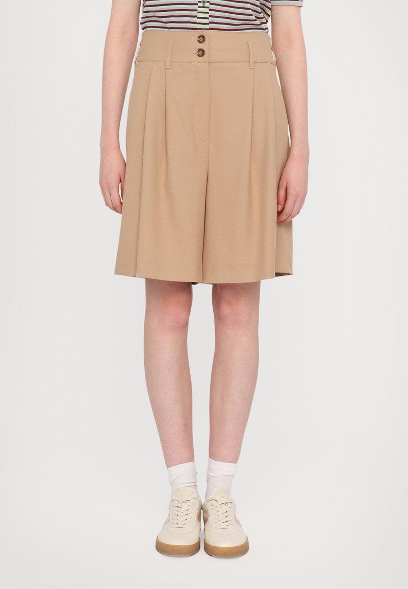 Beige tailored shorts with pleats and two front buttons. Worn with a striped shirt and light-colored shoes, presenting a relaxed fit.