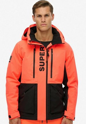 Chaqueta impermeable de un brillante color naranja con una sección inferior negra, destacada marca "SUPER", capucha ajustable y múltiples bolsillos con cremallera.