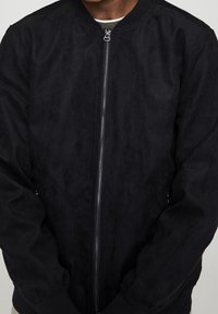 Blouson bombardier noir en daim avec fermeture éclair avant, poignets et col côtelés, doté de deux poches latérales et d'une texture lisse.