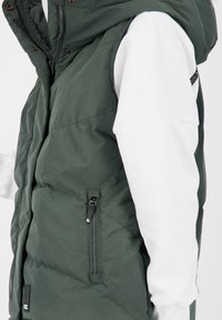 Gilet verde imbottito con cappuccio, caratterizzato da un design trapuntato diagonale e una tasca con zip sul lato sinistro. Indossato sopra un top bianco a maniche lunghe.