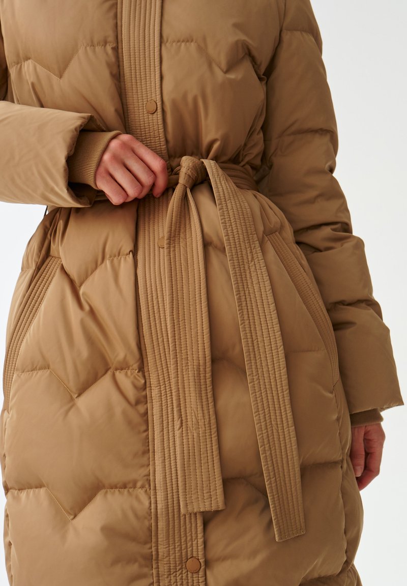 Beżowy ocieplany płaszcz z teksturowanym wzorem diamentowym, z paskiem w formie ściągacza, zapięciem na guziki i dzianinowymi mankietami. Materiał zewnętrzny jest gładki i lekki.