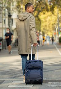 Maleta de navy con ruedas, con múltiples bolsillos con cremallera, fabricada en tela resistente y con un mango retráctil, siendo arrastrada por una persona con un abrigo beige.