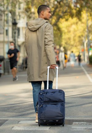 Wheeled navy suitcase with multiple zippered pockets, durable fabric, and retractable handle, being pulled by a person in a beige coat.
