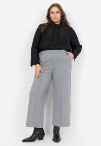 Svart skjortblus med volangdetaljer, i kombination med ljusgrå vida byxor med vertikala sömmar. Svarta ankelskor fulländar looken.
