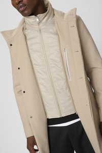 Abrigo de lana beige con una chaqueta interior acolchada desmontable de un beige claro, cierre con cremallera y costuras tonales. La chaqueta interior tiene un cuello estructurado.