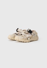Zapatos ligeros de exterior en color beige con parte superior de malla, tiras ajustables en marrón y negro, suelas de goma planas con patrones de tracción.