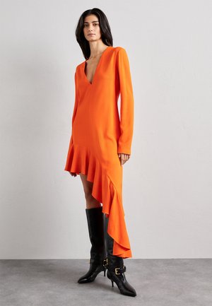 Pomarańczowa sukienka z długim rękawem, głębokim dekoltem w serek i asymetrycznym falbaniastym dołem. Połączona z czarnymi skórzanymi kozakami do kolan z złotymi akcentami.