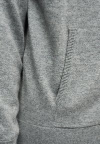 Tissu de pull en maille grise avec une grande poche avant, un ourlet et des poignets côtelés, présentant une apparence douce et texturée.