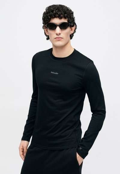 Black long-sleeve shirt with fitted design and a round neckline. Subtle logo on the chest. Model wearing sleek black sunglasses.