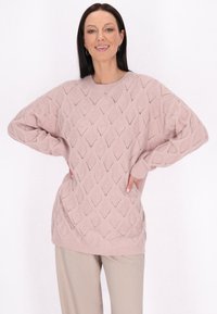 Helles rosa gestricktes Sweatshirt mit Rautenmuster, oversized Schnitt, gerippte Bündchen und Saum; kombiniert mit beigen Hosen.