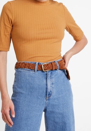 Mujer con una camiseta mostaza de manga corta con canalé metida en unos vaqueros de tiro alto azul claro con un cinturón trenzado de cuero marrón.