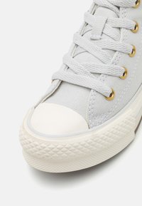 Zapatilla de lona gris con puntera redonda de goma blanca, entresuela texturizada y ojales dorados, presenta cordones planos grises entrelazados en los ojales.