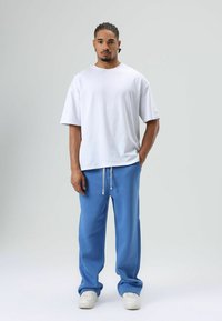 Witte katoenen T-shirt met korte mouwen, gecombineerd met blauwe joggingbroek met een trekkoord. De achtergrond is lichtgrijs, waardoor de outfit centraal staat.