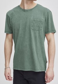 Camiseta de algodón verde con cuello redondo, mangas cortas y un bolsillo en el pecho que presenta un sutil texto en relieve; acabado suave y ligeramente desgastado.