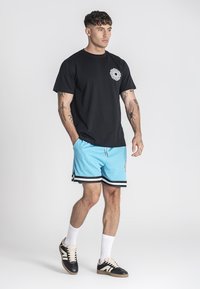 Joven con una camiseta gráfica negra, pantalones cortos deportivos azules con rayas negras y blancas, calcetas blancas y zapatillas negras, de pie contra un fondo blanco.