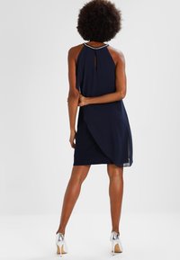 Robe halter marine avec un dos en forme de trou de serrure, design drapé et détails en ourlet transparent. Accentuée d'un col perlé, associée à des talons hauts argentés.