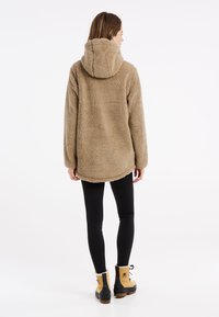 Beige, nadýchaný fleecový svetr s kapucí, volný střih, kulatý lem a dlouhé rukávy. Nosený s černými legínami a hnědými botami.