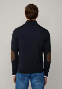 Marinblå tröja med hög krage och bruna armbågsfickor, strukturerad stickad material och ribbade manschetter, kombinerad med blå jeans.