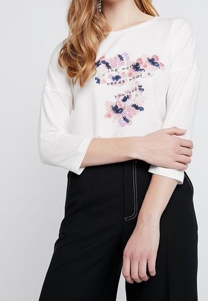 Vrouw die een wit overhemd met lange mouwen draagt met roze en paarse bloemen en de tekst "The world needs what you got,", gecombineerd met een zwarte broek.
