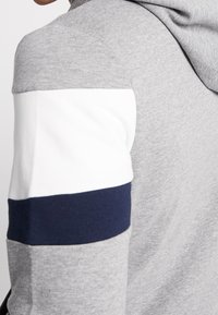 Sudadera con capucha gris de textura suave, con rayas horizontales blancas y azul marino en las mangas. Diseñada con un ajuste holgado.