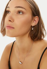 Orecchini a forma di goccia in argento e collana coordinata con una finitura liscia e lucida, posti su uno sfondo chiaro e indossati con una maglietta nera.