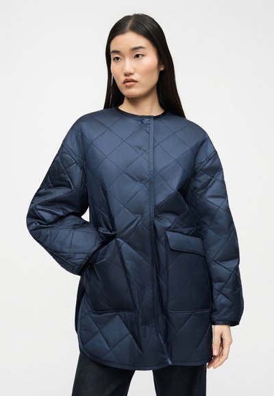 Γυναίκα με μακριά σκούρα μαλλιά που φοράει μπλε-σκούρο μπουφάν με μεγάλα τσεπάκια, στέκεται με το ένα χέρι στην τσέπη μπροστά σε απλό φόντο.