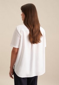 Camicia bianca a maniche corte con una vestibilità rilassata e orlo arrotondato. Tessuto liscio. La vista posteriore mostra capelli lunghi e dritti di colore castano.