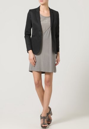 Femme portant une robe grise jusqu'aux genoux, un blazer noir cintré et des sandales grises à talons hauts, debout devant un fond uni.