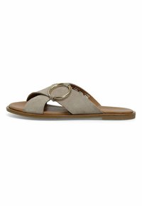 Sandalia de ante tipo slip-on en beige, con diseño de bandas cruzadas, un acento circular dorado y una suela plana marrón. Textura suave.