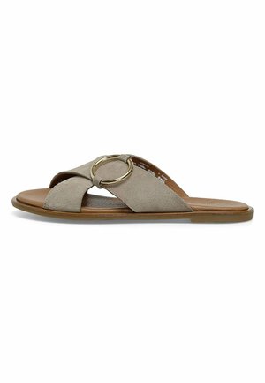 Sandalia de ante tipo slip-on en beige, con diseño de bandas cruzadas, un acento circular dorado y una suela plana marrón. Textura suave.