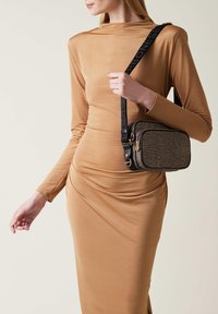 Black and gold bag with textured surface and zip closure, featuring a black strap with logo. Worn at the hip against a fitted beige dress.