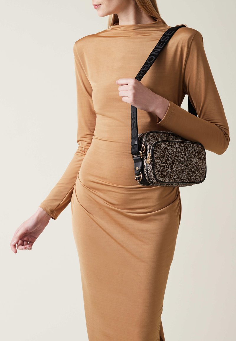 Black and gold bag with textured surface and zip closure, featuring a black strap with logo. Worn at the hip against a fitted beige dress.