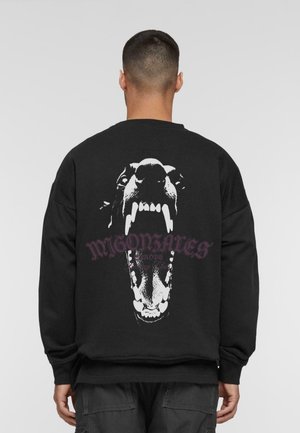 Mężczyzna w czarnej bluzie z dużym białym nadrukiem przedstawiającym ryczącą twarz psa oraz stylizowanym napisem na plecach.