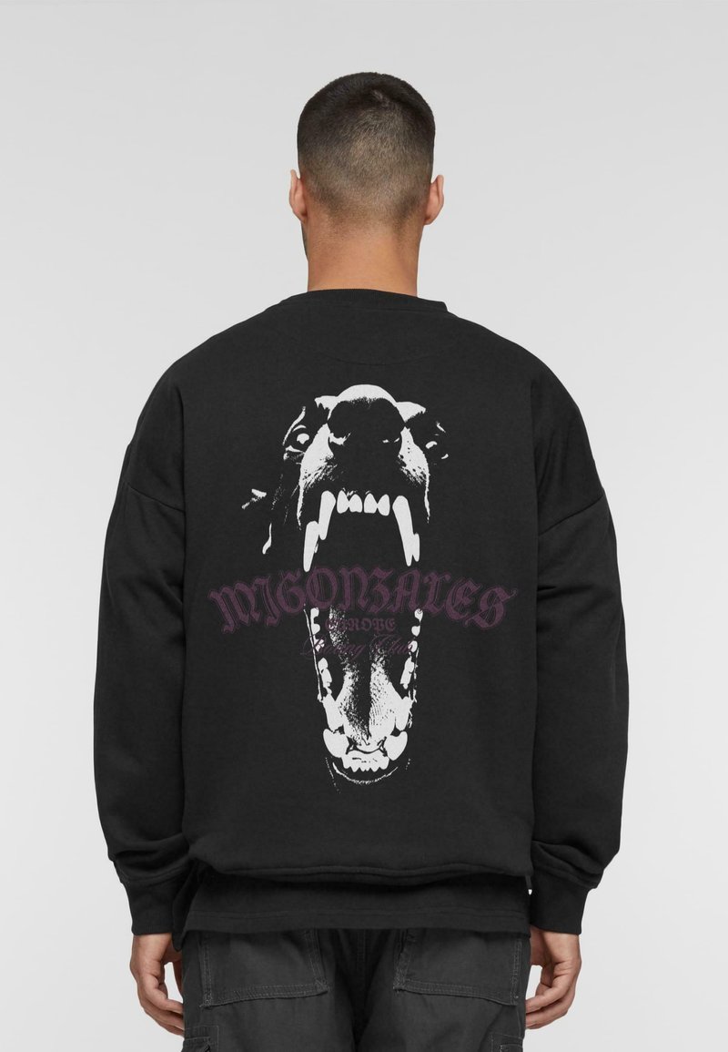 Mężczyzna w czarnej bluzie z dużym białym nadrukiem przedstawiającym ryczącą twarz psa oraz stylizowanym napisem na plecach.