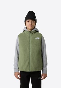 Grön fleeceväst med hög krage, full framdragkedja och logotyp på vänster bröst, bärs över en grå hoodie. Modellen har en svart mössa.