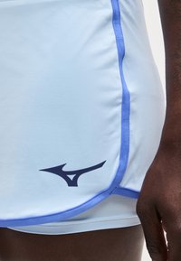 Κοντινή λήψη ανοιχτό μπλε αθλητικών σορτς με μωβ διακόσμηση και μαύρο λογότυπο Mizuno κοντά στο τελείωμα, φορεμένα από άτομο με σκουρόχρωμο δέρμα.