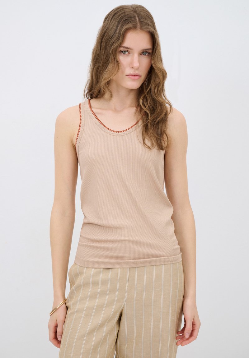 Beige ribbet tanktop med rød syning langs halsudskæringen. Bærer let lysebrune stribede bukser. Glat tekstur og tætsiddende design.