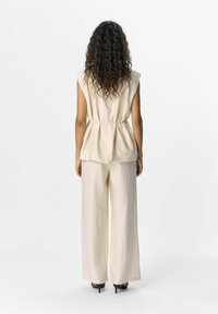 Top beige senza maniche con vita arricciata, abbinato a pantaloni beige a gamba larga, con una texture morbida e una silhouette rilassata.
