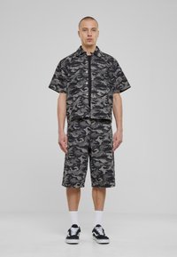 Kortermet skjorte og shorts med camo-mønster i gråtoner, laget av vevd stoff. Kombinert med svarte joggesko og hvite sokker.