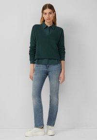 Dunkelgrüner strukturierter Pullover mit V-Ausschnitt und Rippdetails, über ein Hemd mit Kragen getragen, kombiniert mit hellblauen Jeans und weißen Sneakern.