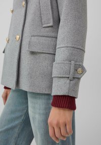 Veste croisée en mélange de laine grise avec boutons dorés, épaules structurées, poches à rabat et détails côtelés aux poignets burgundy.