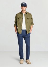 Olivgrünes Hemd mit Knöpfen und zwei Brusttaschen, darunter ein weißes T-Shirt, marineblaue Hose und beiges Schuhwerk, kombiniert mit einer marineblauen Kappe mit weißem Schriftzug.